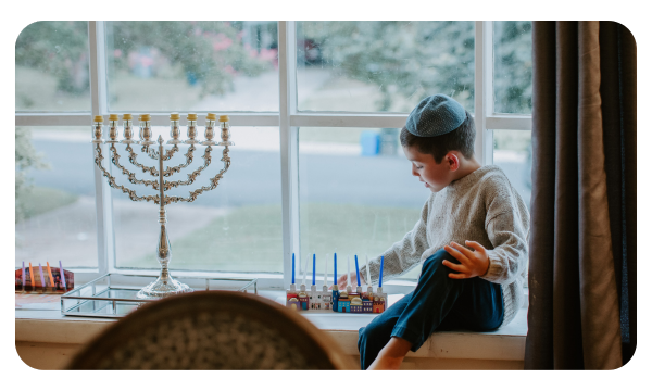 Kid with a yamulka sitting by the window and putting candles into a menorah